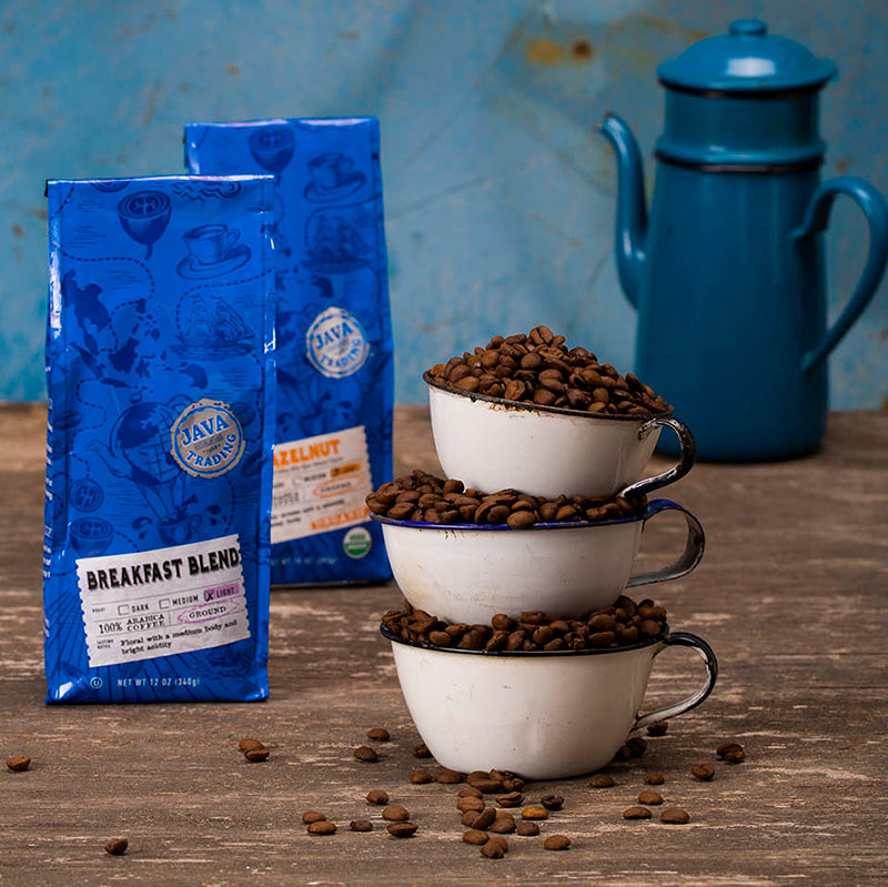 two blue bags of java trading and three piled mugs with roasted beans